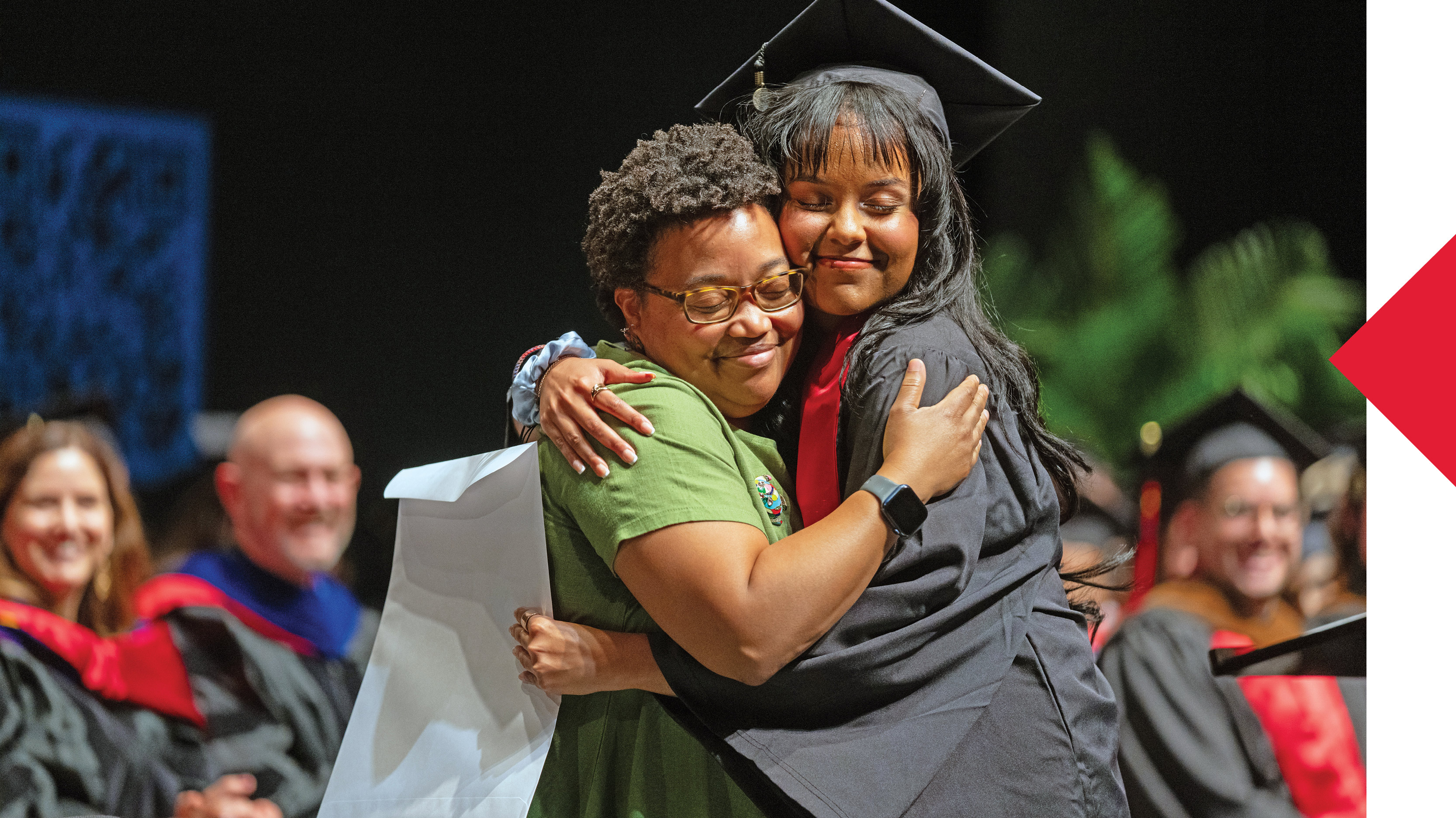 ull-color photograph of a joyous graduation moment. A faculty member in green embraces a graduating student in cap and gown, both smiling with eyes closed in pride and connection. The background shows other graduates and faculty in regalia, symbolizing achievement and community.