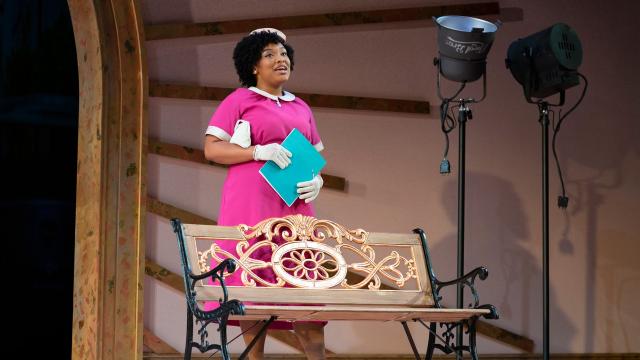 A Black woman holds a blue folder and stands behind a bench on stage.